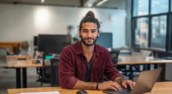 Junger Mann mit Dutt am Laptop, lächelnd. Ein zukünftiger marketing master hamburg.