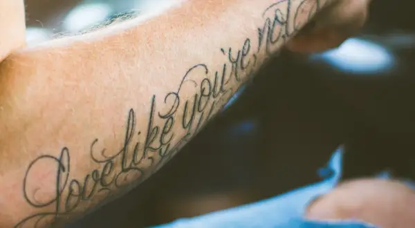 Ein Arm mit einem geschwungenen Schriften Tattoo, das die Worte "Love like you're not" trägt.