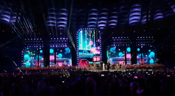 Wydarzenia na Stadionie Narodowym: scena z neonowymi samochodami i tańczącymi artystami, oświetlona przez reflektory. Tłum widzów wpatrzony w widowisko.