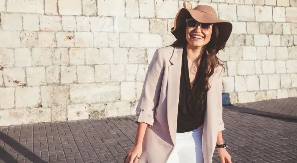 Donna sorridente con cappello e occhiali da sole, perfetta per come vestirsi con 20 gradi: blazer leggero, top nero e pantaloni bianchi.