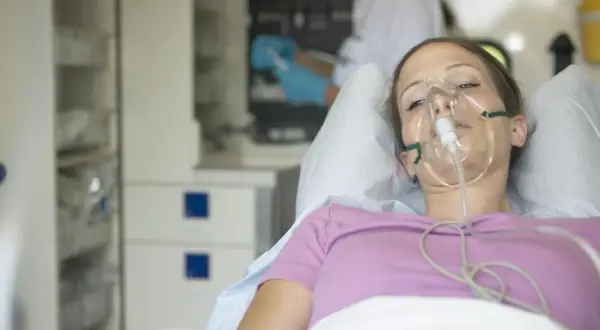 Młoda kobieta w masce tlenowej, leżąca w łóżku szpitalnym. Jej stan sugeruje chorobę popromienną, wymagającą intensywnej opieki medycznej.