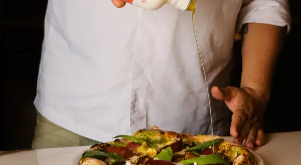 Un cuoco versa l'olio Coratina migliore su una pizza gourmet. L'olio extra vergine d'oliva esalta i sapori.
