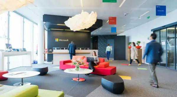 Un **open space** moderne chez Microsoft. Une femme travaille sur son ordinateur portable, entourée de canapés colorés. Des affiches "Imagine", "Achieve", "Innovate" flottent au plafond.
