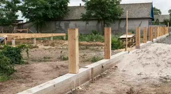Solidny fundament pod ogrodzenie z betonowym murem i drewnianymi słupkami. W tle widać budowę domu.