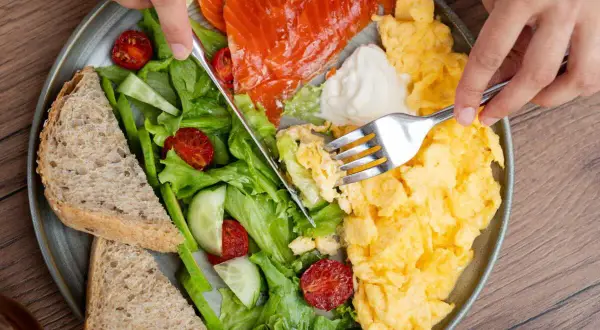 Assiette colorée avec saumon fumé, œufs brouillés, salade verte, tomates séchées, concombre et pain complet. Un repas protéiné parfait pour bien commencer la journée.