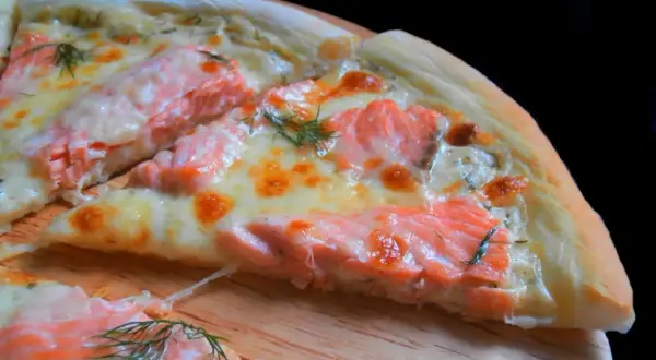Kawałek pysznej pizzy z łososiem, roztopionym serem i koperkiem na drewnianej desce.