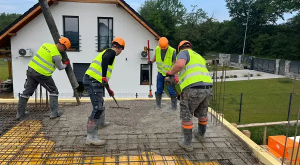 Budowlańcy w kamizelkach i kaskach, przestrzegając zasady bhp, wylewają beton. Jeden kieruje rurę, drugi grabi, trzeci wyrównuje.