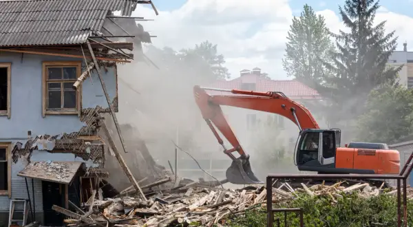 Pomarańczowy koparka prowadzi rozbiórkę budynku, wzbijając tumany kurzu. Ruiny i drewno leżą na ziemi.