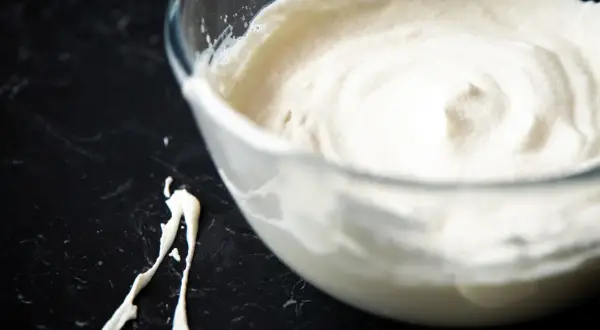 Schlagrahm in einer Glasschüssel, bereit für ein Dessert. Ein Klecks Sahne ist daneben auf der dunklen Arbeitsfläche zu sehen.
