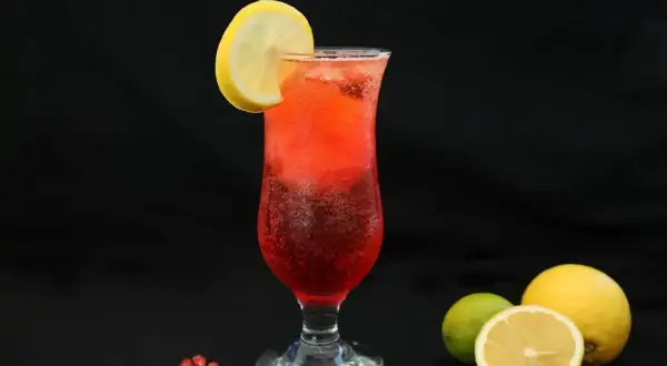 Orzeźwiający drink z sokiem z granatu, ozdobiony plasterkiem cytryny. Obok leżą owoce granatu, cytryny i limonka.