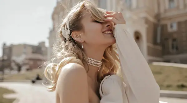 Uśmiechnięta blondynka w perłowej biżuterii, z wpiętymi we włosy prawdziwymi perłami, cieszy się słońcem.