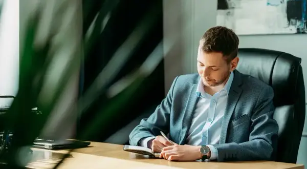 Mężczyzna w garniturze siedzi przy biurku i pisze w notatniku.