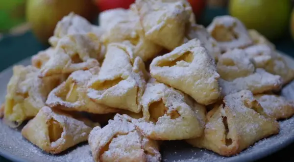 Stos ciasteczek serowych z jabłkami, oprószonych cukrem pudrem, na talerzu. W tle jabłka.