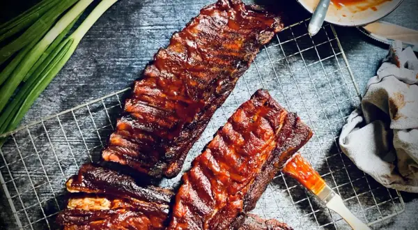 Saftige spare ribs glänzen in BBQ-Sauce auf einem Grillrost. Frühlingszwiebeln und eine Sauce-Schale deuten auf ein köstliches Mahl hin.