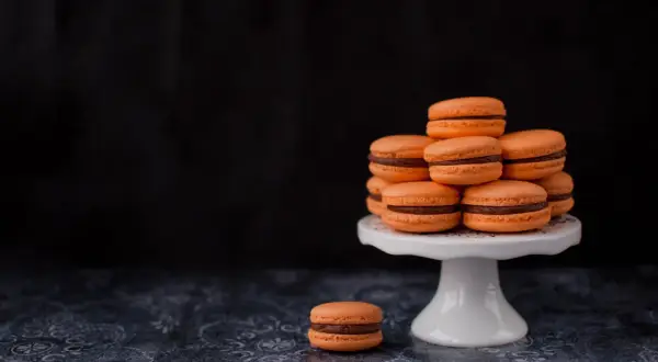 Pomarańczowe makaroniki francuskie z czekoladowym nadzieniem piętrzą się na białej paterze. Jeden makaronik leży obok.