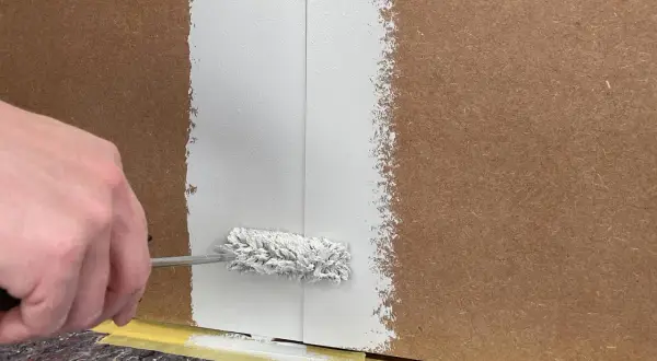 Hand streicht mit einer Farbrolle weiße Farbe auf folierte Paneele. Der Boden ist mit Malervlies und Klebeband geschützt.