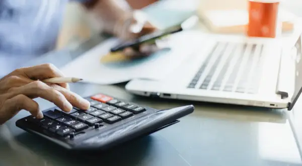 Dłoń z ołówkiem liczy na kalkulatorze, obok laptop i kubek. Analiza rachunków, może zużycie kwh.