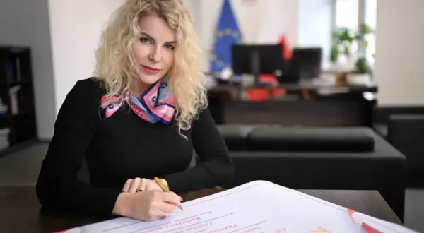 Kobieta z kręconymi blond włosami, w czarnej bluzce i kolorowym szalu, podpisuje dokument. Jest rzecznikiem małych i średnich przedsiębiorców.