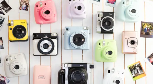 Różne rodzaje aparatów fotograficznych Instax w jaskrawych kolorach, ułożone na białym tle, otoczone odbitkami zdjęć.