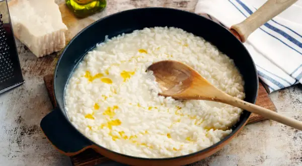 Krämig parmesanrisotto i en panna, med en träsked som rör om. Ingredienser som parmesanost och olivolja syns i bakgrunden.