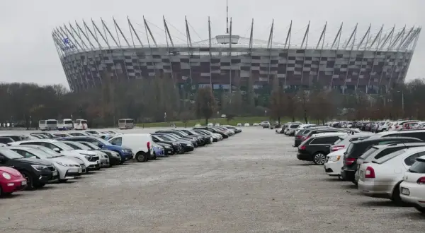 Duży parking przed Stadionem Narodowym w Warszawie. Widać rzędy samochodów i autobusów na błoniach stadionu.