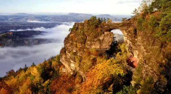 Majestätische Felsformation Pravcicka brana mit einem natürlichen Torbogen, umgeben von herbstlichem Laub und Nebel im Tal.