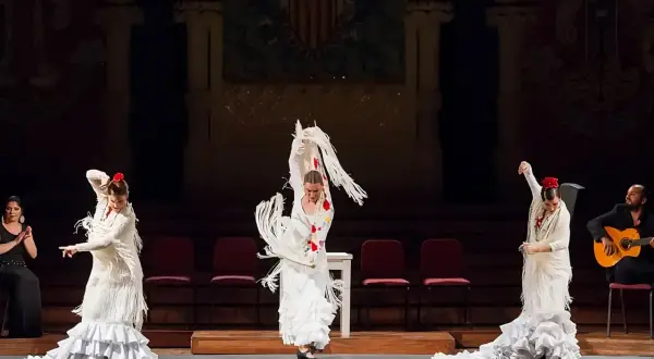 Mujeres en un apasionado baile flamenco, con trajes blancos y volantes, acompañadas por un guitarrista.