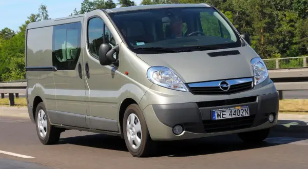 Srebrny Opel Vivaro 2.0 CDTI z przyciemnianymi szybami jedzie po drodze, otoczony zielenią drzew.
