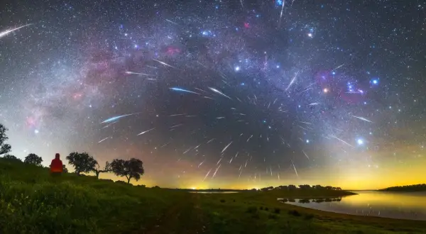 A lone figure watches a spectacular display of shoot stars streaking across the Milky Way above a tranquil lake and grassy hills.