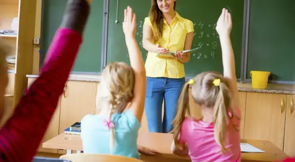 Nauczycielka w żółtej bluzce stoi przed tablicą, a uczniowie podnoszą ręce.