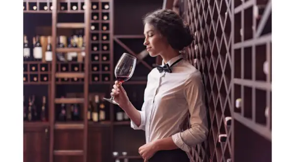 Elegancka sommelierka w białej koszuli i muszce wącha czerwone wino w kieliszku, otoczona regałami pełnymi butelek.