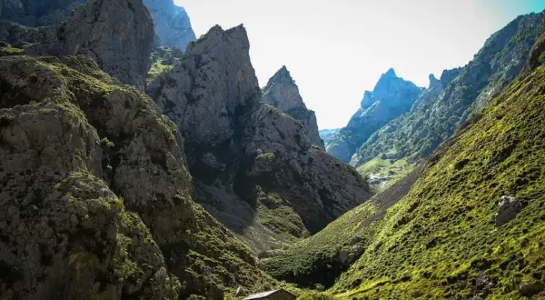 Kamienna chata w dolinie u stóp skalistych szczytów, przypominających kształtem **Naranjo de Bulnes**.