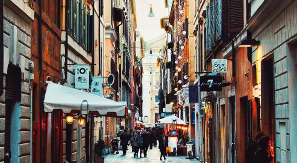 En livlig gränd i ett kvarter i Rom, med butiker och restauranger längs sidorna. Folk promenerar och njuter av atmosfären.