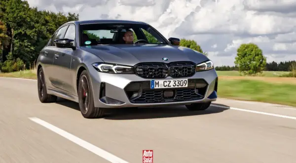 Szare BMW 320d jedzie po drodze, za kierownicą siedzi mężczyzna. W tle drzewa i chmury.