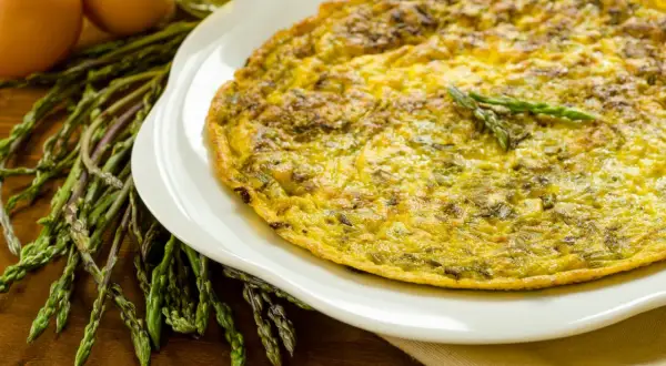 Frittata di asparagi selvatici dorata su piatto bianco, con asparagi freschi e uova sullo sfondo.