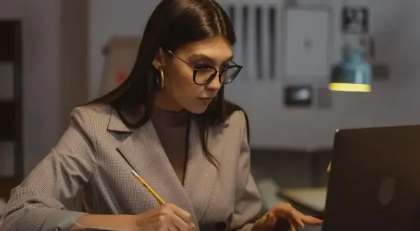 Kobieta w okularach pracuje przy laptopie, notując w zeszycie. Zastanawia się nad limitem nadgodzin w roku, by zachować równowagę.