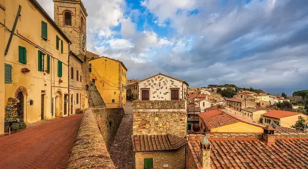Un vicolo con case gialle e un campanile a Montescudaio. Tetti rossi e cielo nuvoloso, un'atmosfera suggestiva per scoprire cosa vedere.