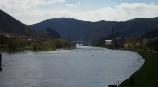 Flusslandschaft bei Ústí nad Labem mit bewaldeten Hügeln, Industriegebäuden und einer Stadt am Ufer.