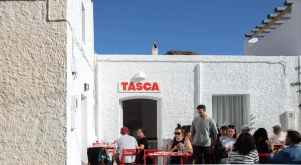 Gente disfrutando en el bar Tasca, con sus altas hortichuelas, bajo un cielo azul.