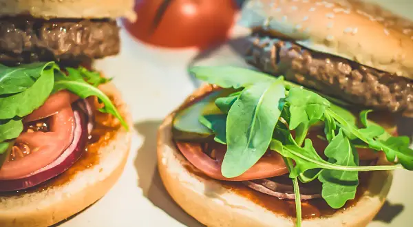 Dwa domowe hamburgery z bułką, kotletem, pomidorem, czerwoną cebulą i rukolą.
