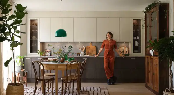 En kvinna i orange jumpsuit lutar sig mot köksbänken i ett kök med fransk inredningsstil. Ett runt träbord med stolar står framför köksöarna.