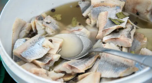 Kawałki śledzi w zalewie z przyprawami, gotowe do podania. Widać łyżkę zanurzoną w marynacie.