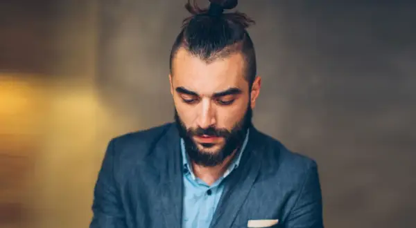Mężczyzna z brodą i modną fryzurą samuraj, w eleganckim garniturze, spogląda w dół.