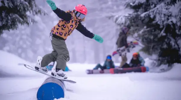 Dziecko na snowboardzie balansuje na rurze w spindleruv mlyn snowpark. W tle inni snowboardziści.