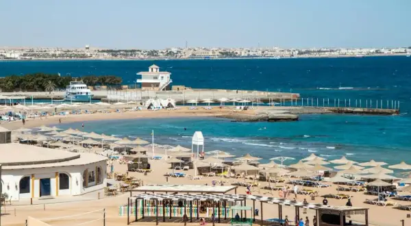 Solig strand i Hurghada, Egypten, med parasoller, solstolar och turkost vatten. En båt ligger vid en pir.
