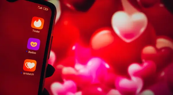 Na ekranie telefonu widać ikony aplikacji randkowych: Tinder, Badoo i W-Match. Tło to czerwone serca, symbolizujące miłość i poszukiwanie tej jedynej osoby. Wybór bezpiecznych portali randkowych to pierwszy krok do znalezienia szczęścia.