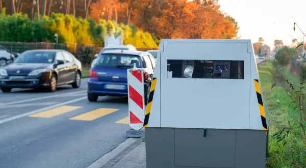 Un radar de chantier surveille la circulation sur une route bordée d'arbres aux couleurs d'automne. Des voitures passent devant.