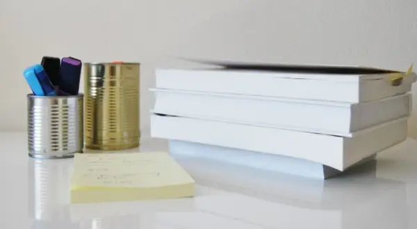 Na biurku stos białych książek, obok puszki z pisakami i żółta karteczka z notatkami, które mogą posłużyć jako akapity w rozprawce.