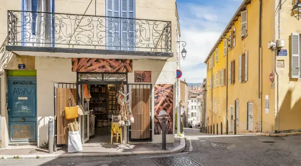 Bunte Häuser und ein Gewürzladen in der **Marseille Altstadt**. Eine schmale Gasse führt bergauf, gesäumt von sonnengelbem Mauerwerk und blauen Fensterläden.