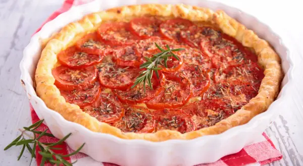 Une délicieuse tarte à la tomate, garnie de fines herbes et d'une branche de romarin frais, repose dans un plat blanc sur un torchon à carreaux rouges.